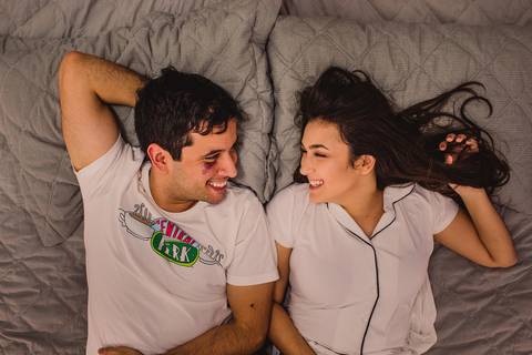 Casal deitando na cama. Olhando um para o outro e sorrindo, após guerrinha de travesseiro. Ensaio Fotográfico realizado em Vitória. Bairro: Enseada do Suá. Estado: Espírito Santo.'