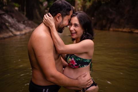 Fotografia da gestante na cachoeira sorrindo junto com seu esposo. Eles estão abraçados. Retrato foi feio no momento em que ela está gargalhando junto com ele.'