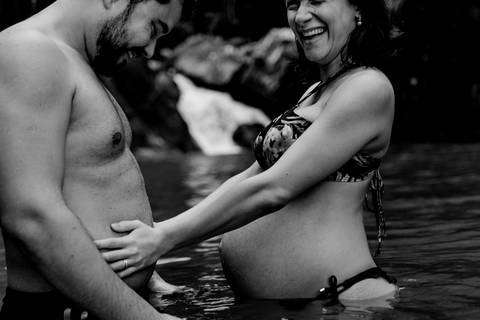 Fotografia preto e brando da gestante segurando a barriga do esposo.'