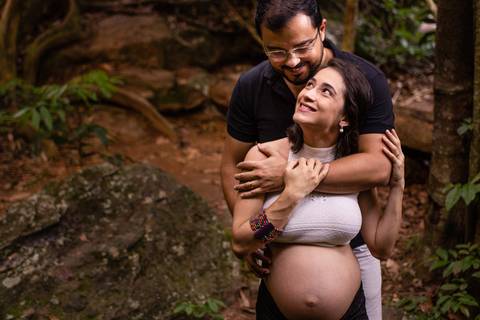 Fotografia de ensaio de gestante. Esposa olhando para esposo. Ele está abraçado por detrás dela e ela sorrindo para ele.'