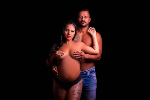 A gestante e seu esposo estão abraçados em um momento íntimo, com as mãos carinhosamente posicionadas na barriga, simbolizando o amor e a cumplicidade do casal nessa jornada da maternidade.'