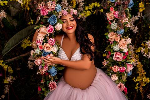 A gestante posa delicadamente, usando um adereço floral no cabelo, evocando uma sensação de feminilidade e conexão com a natureza.'