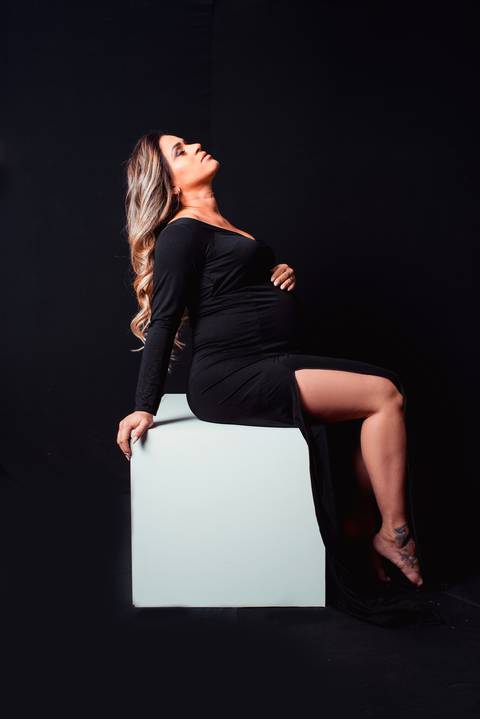 A gestante esbanja confiança e sensualidade nas fotos com vestido preto e fundo preto, criando um contraste marcante e misterioso.'