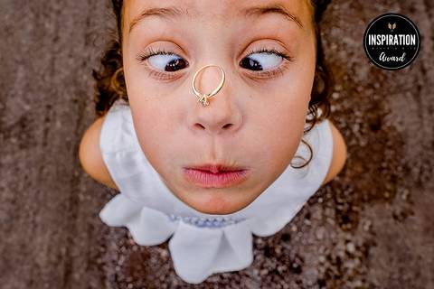 Menina em momento divertido e descontraído equilibrando um anel no nariz. Uma imagem premiada que retrata a espontaneidade e a alegria das crianças em celebrações de casamento, adicionando leveza e humor ao evento.'