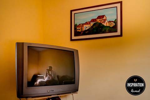 Reflexo de um casal em uma antiga televisão, com uma pintura ao fundo, criando uma imagem que mistura nostalgia e intimidade. Essa fotografia premiada destaca a conexão entre tradição e amor, em um ambiente aconchegante e familiar.'