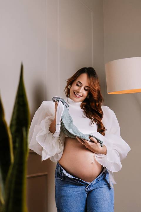 Carol vivendo um instante carinhoso ao segurar a roupinha do bebê durante o ensaio de gestante no estúdio.'