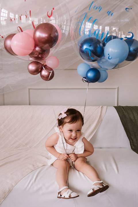 Menina sorrindo sentada na cama com balões rosa e azul ao fundo'