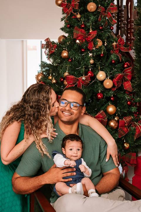 Mãe beijando o rosto do pai enquanto seguram o bebê diante da árvore'