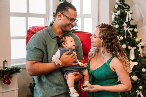 Família posicionada em frente à árvore de Natal sorrindo com o bebê'