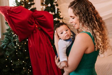 Mãe abraçando o bebê ao lado da árvore decorada'