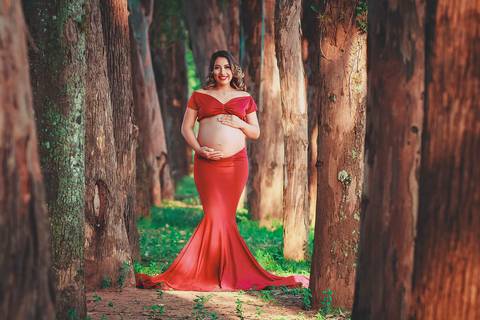 gestante com vestido vermelho e caminho de árvores, encarando a câmera, no Parque dos Ipes, em Santa Bárbara d'Oeste - SP'