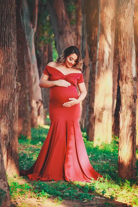 gestante fazendo carinho em sua barriga e sorrindo, com vestido vermelho em um caminho de árvores, no Parque dos Ipes, em Santa Bárbara d'Oeste - SP'