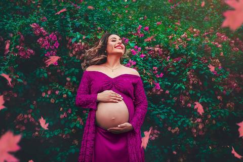 gestante jogando o cabelo para os lados num lugar bem florido e com as mãos na barriga de vestido rosa, no Parque dos Ipes, em Santa Bárbara d'Oeste - SP'