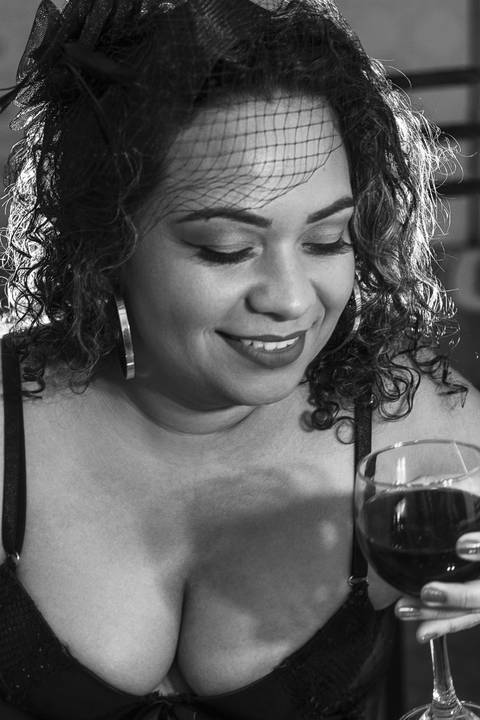 Fotografia de ensaio sensual boudoir plus size de mulher morena de cabelo cacheado vestida com lingerie preta tomando vinho'