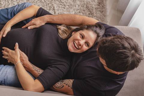 Marido e mulher grávida sorrindo deitados no sofá de casa '