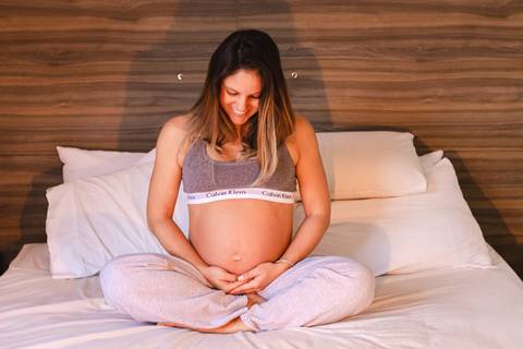 mulher grávida sentada na cama abraçando a barriga e sorrindo '