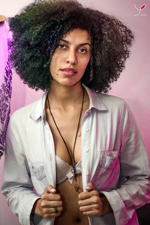 Foto sensual da modelo negra com jaqueta jeans e sutiã, cabelos crespos e longos. Num local com uma luz rosa, diferenciada.'