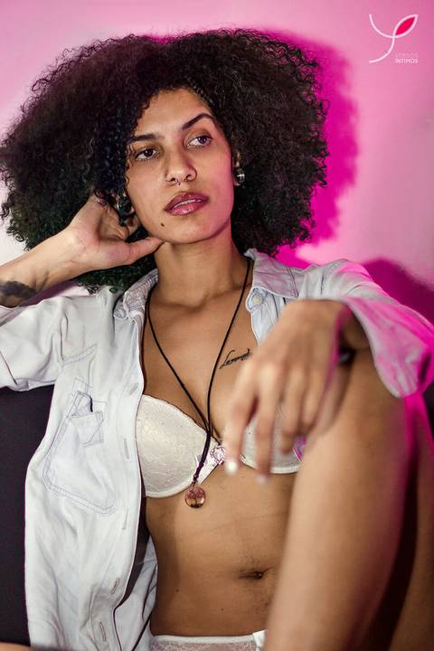 Foto sensual da modelo negra com jaqueta jeans e sutiã, cabelos crespos e longos. Num local com uma luz rosa, diferenciada.'