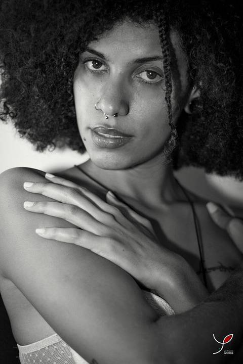 Foto sensual da modelo negra apenas com sutiã, ela tem cabelos crespos e longos. Numa foto em preto e branco, com ela se abraçando.'