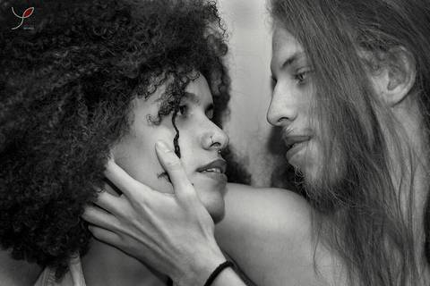 Casal olhando um para o outro. Ele sem camiseta e ela num estilo sensual. Enquanto o namorado faz carinho no rosto da namorada - que é negra de cabelos crespos.'