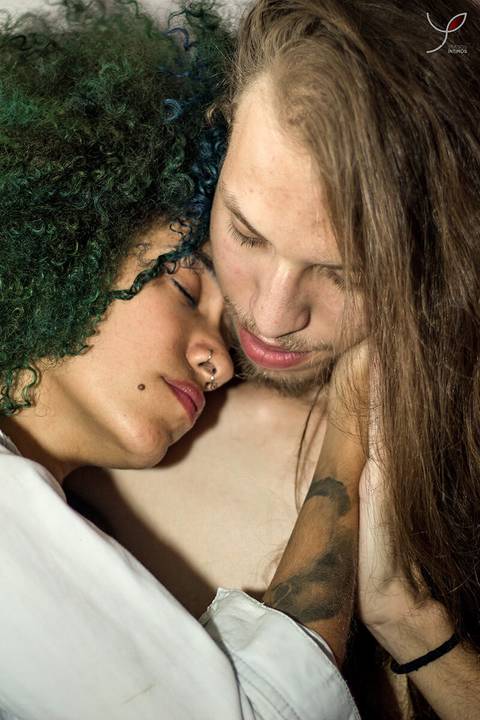 Casal olhando um para o outro. Ele sem camiseta e ela num estilo sensual. A modelo negra está deitada junto com o namorado, num momento de carinho.'