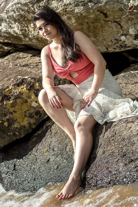 A modelo está sentada numa pedra próxima a cachoeira. Usando um vestido branco e uma camisa rosa.'