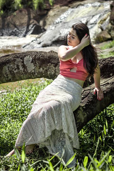 A modelo está sentada num tronco próximo a cachoeira. Usando um vestido branco e uma camisa rosa.'