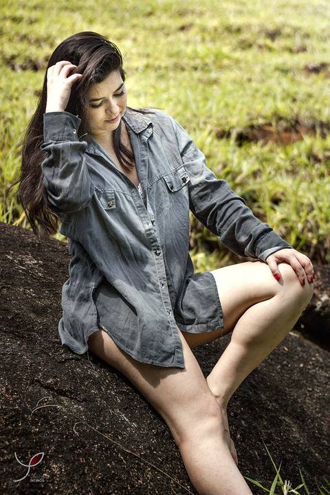 A modelo está sentada numa pedra próxima a cachoeira. Usando uma camisa longa sobre o sutiã - e sorrindo.'