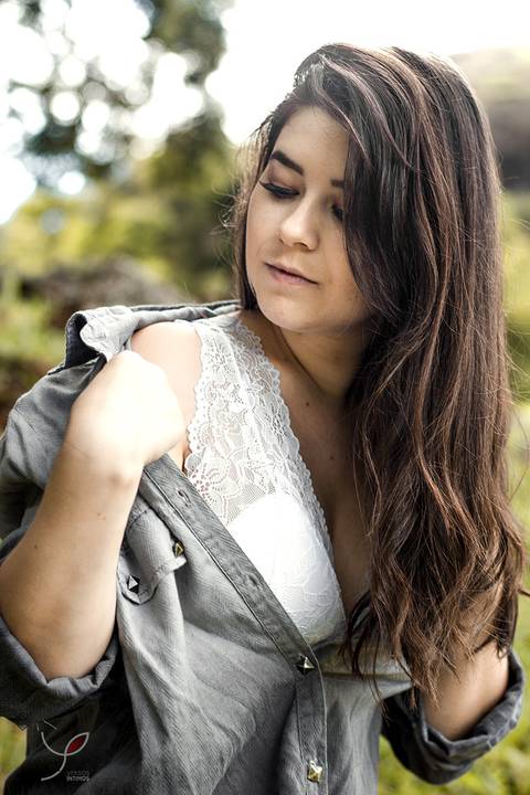 A modelo está sentada numa pedra próxima a cachoeira. Usando uma camisa longa sobre o sutiã - e sorrindo. E puxando o lado da camisa, mostrando a roupa íntima.'
