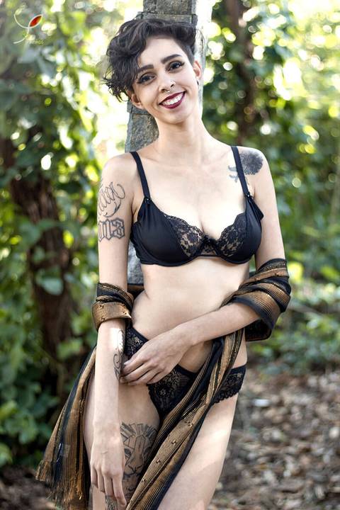 Nesta foto sensual a modelo está com um sutiã preto e um chale marrom, num campo e olhando séria para o fotógrafo.'