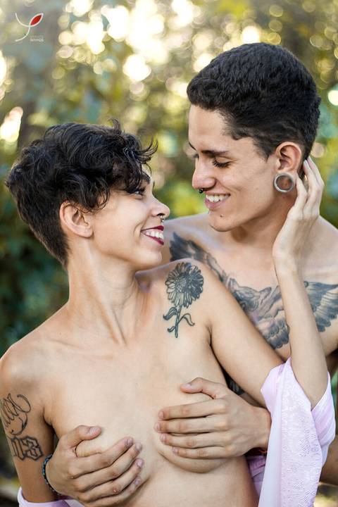 A modelo e o namorado - o casal, estão de topless e olhando um para o outro sorrindo. Ambos tem tatuagem. No cenário vemos a natureza atrás. '
