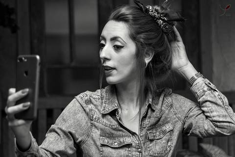 Fotografia em preto e branco da modelo olhando para o celular enquanto arruma o cabelo. Ela está usando jaqueta jeans e cabelo preso.'