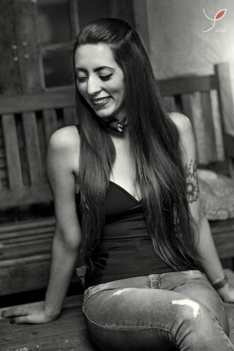 Fotografia em preto e branco da modelo sentada na mesa de uma cafeteria. Ela veste uma camisa preta e tem tatuagem nos braços. Na imagem ela olha para o lado.'