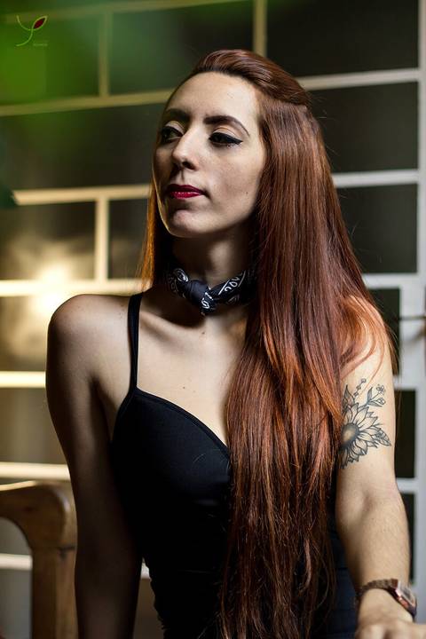 Modelo ruiva com uma camisa preta, laço no pescoço, tatuagem no braço sentada numa mesa e olhando para o lado. Numa cafeteria de São José dos Campos.'