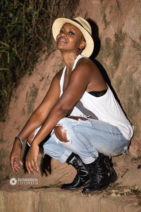 Modelo negra está numa escadaria de noite, de camiseta regata branca e chapéu. Ela está sentada de lado. Numa ensaio fotográfico urbano.'