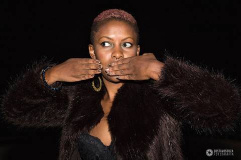 Modelo negra num ensaio noturno urbano. Ela está de roupas pretas, blusa de pêlos e fazendo um sinal de beijo.'