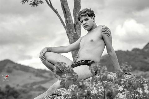 O modelo branco, de cueca e sem camiseta, está sentado numa pedra e olhando para a câmera. Numa fotografia em preto e branco com a natureza no fundo.'