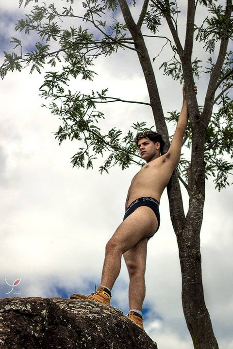 O modelo branco, de cueca e sem camiseta, está encostado numa árvore.'