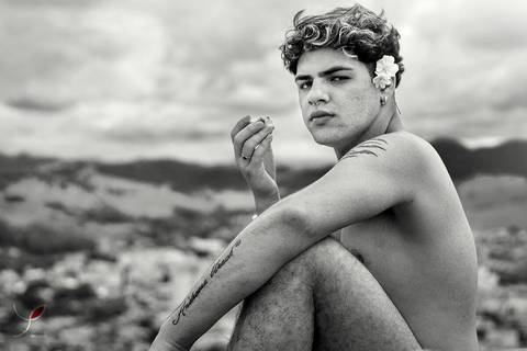 O modelo branco, de cueca e sem camiseta, está sentado numa pedra e olhando para a câmera. Com a cidade no fundo e numa foto em preto e branco.'