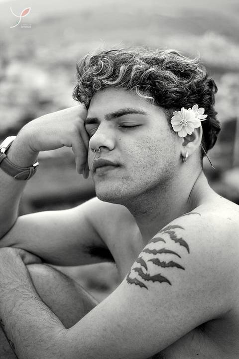 O modelo branco, de cueca e sem camiseta, está sentado numa pedra e de olhos fechados. Com a cidade no fundo.'