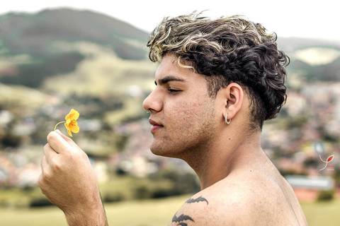 O modelo branco, de cueca e sem camiseta, está sentado numa pedra e está segundo uma flor. Com a cidade no fundo.'