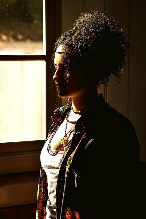 Modelo numa sala de madeira, numa foto artística com uso de sombras. Usando camisa colorida e acessórios hippie'