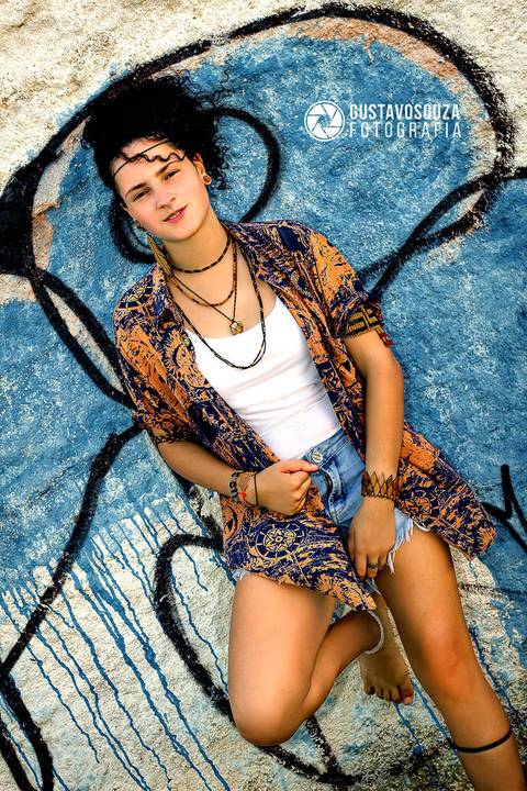 Modelo de cabelo preso, pele clara, jovem, com roupas coloridas em pé e olhando para a câmera, encostada na parede.'