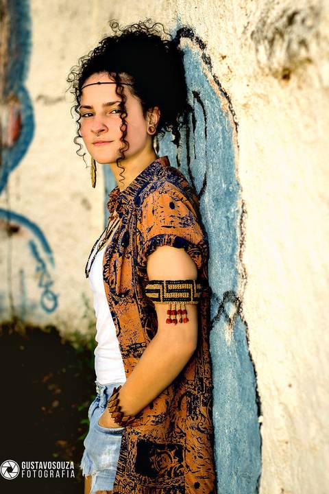 Modelo de cabelo preso, pele clara, jovem, com roupas coloridas em pé e olhando para a câmera, encostada num muro com arte em grafite.'
