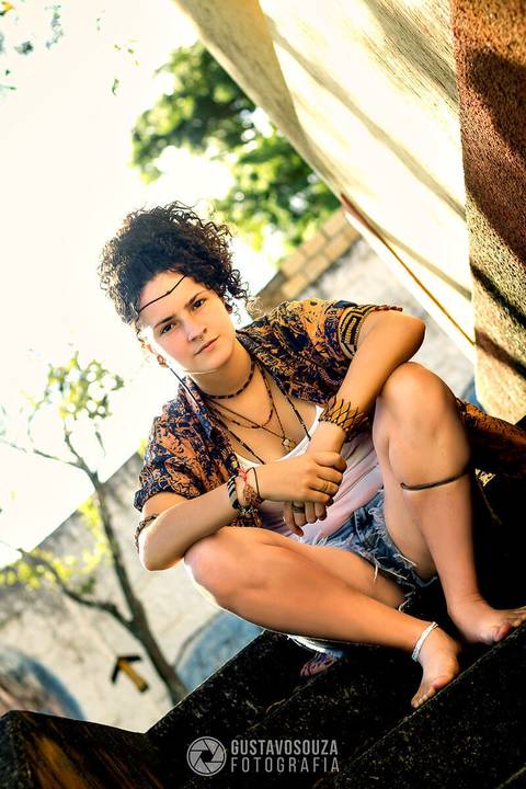 Modelo na área externa do ambiente, sentada a escada, numa pose firme, e um céu azul atrás.'