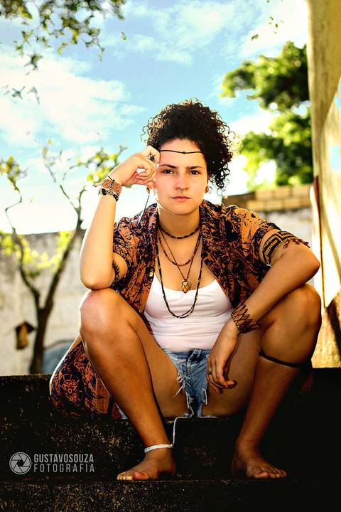 Modelo na área externa do ambiente, sentada a escada, numa pose firme, e um céu azul atrás.'