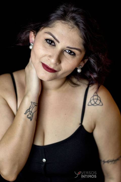 Modelo com tatuagens e vestido preto posando em uma fotografia de estúdio. Ela olha para a câmera.'