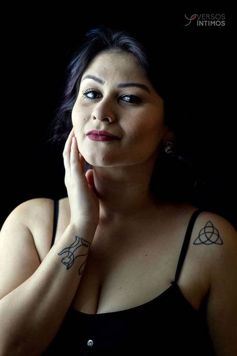 Modelo com tatuagens e vestido preto posando em uma fotografia de estúdio. Ela olha para a câmera.'