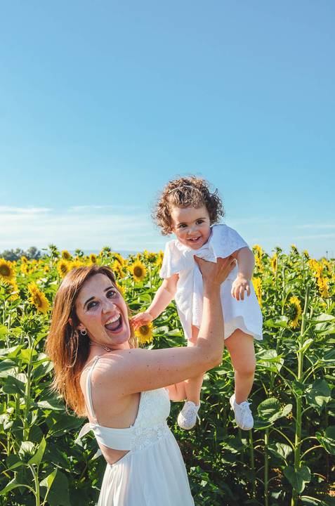 Mulher adulta com criança em sessão familiar brincando em campo de girassóis ao ar livre. Mãe e filha em momento de conexão emocional em ambiente rural natural. Fotografia profissional capturando vínculo familiar e alegria em composição artística.'