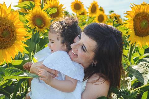 Mulher adulta com criança pequena segurando girassol em sessão familiar. Mãe e filha em momento de conexão natural em campo ao ar livre. Fotografia profissional capturando vínculo emocional familiar em ambiente rural paisagístico.'